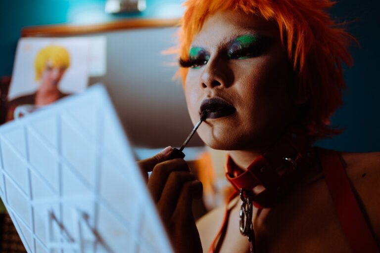 Low angle of crop ethnic eccentric transgender guy with bright makeup in wig and leather collar applying dark lipstick while preparing for performance in nightclub